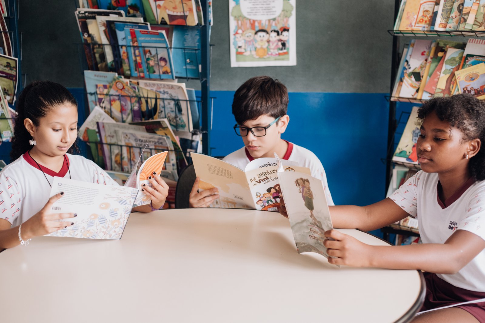 Três alunos dos Anos Iniciais lendo juntos numa mesa redonda da biblioteca do Colégio Goyases